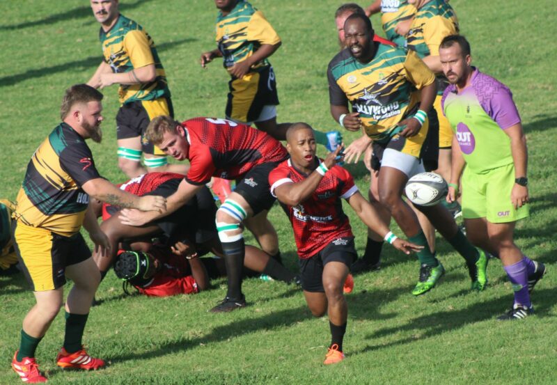 A photo taken of two rival rugby teams during a match on a rugby field. A referee is standing in front of the players.
