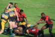 A photo taken of two rival rugby teams during a match on a rugby field. A referee is standing in front of the players.