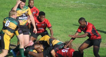 A photo taken of two rival rugby teams during a match on a rugby field. A referee is standing in front of the players.