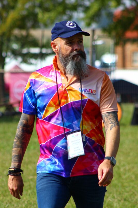 A man dressed in a colourful T-Shirt, sporting a grey beard and tattoos is photographed walking on a field.