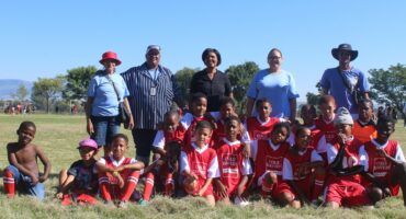 A group of soccer players and event organisers pose for a photo on the soccer field outside in the sun.