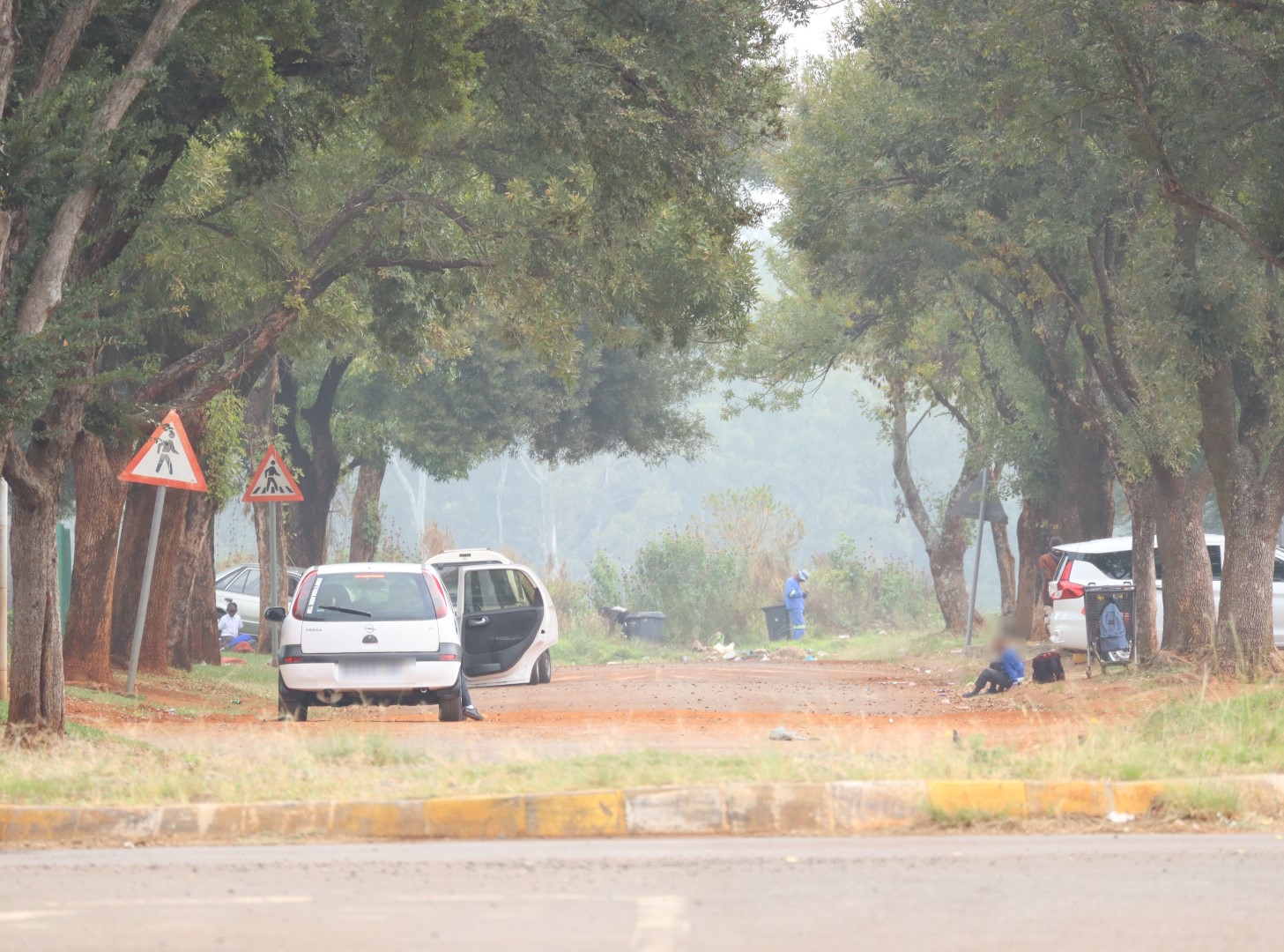 A photo of a road. in the photo is massive overgrown trees, litter and people standing around. The photo also shows a badly deteriorated road.