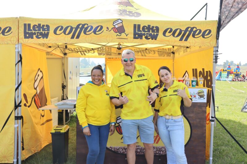 Three people dressed in yellow tshirts and denim pants pose in front of a yellow gazebo selling coffee.