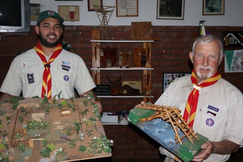 Two Scouts leaders are posing with camp-site models in their hands. They are wearing the full Scouts uniform. Memorabilia is visible in the background.
