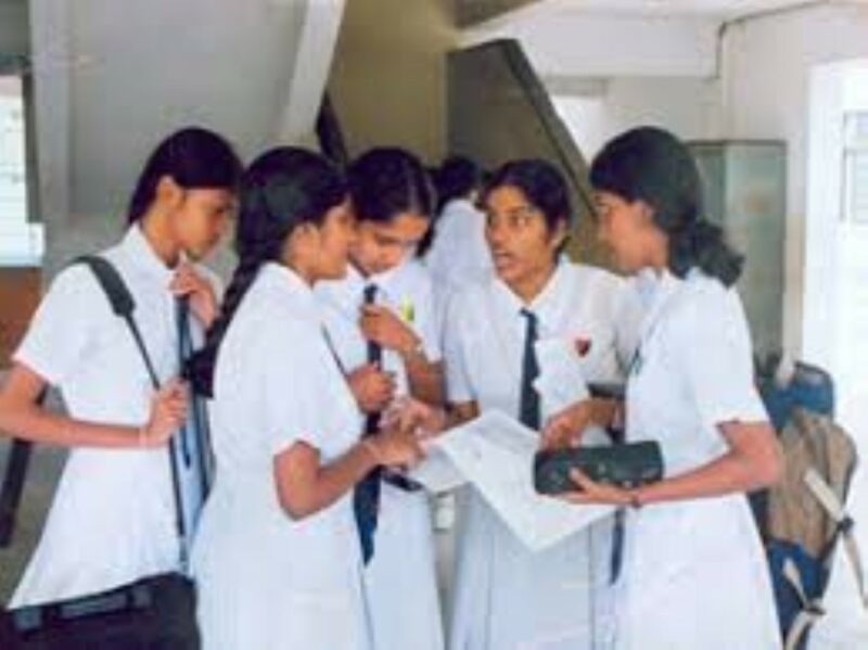 School girls in their all-white school uniforms in a group speaking to each other. They are inside a building. Some students have school related material in their hands.