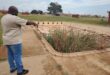 A man dressed in a cream coloured shirt and blue jeans points to an abandoned public swimming pool.