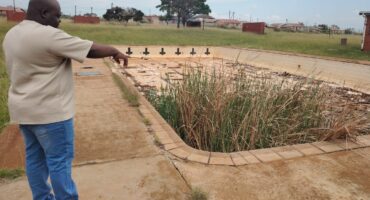 A man dressed in a cream coloured shirt and blue jeans points to an abandoned public swimming pool.