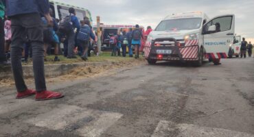 A photo captures several ambulance fleets at an accident scene, where multiple school children from multiple schools were injured. school children and paramedics are seen standing outside around the fleets. In the foreground is a man standing in overalls, red socks and flipflops.