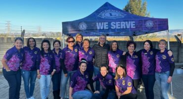 A group of woman dressed in purple pose for a photo. All of them are posed in front of an old, white building.