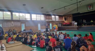 Image of group of wrestlers sitting on the mats while coaches speak to them.