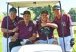 A group of men dressed in maroon shirts and white and black pants sitting inside a golf cart on the golf coarse.