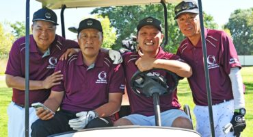 A group of men dressed in maroon shirts and white and black pants sitting inside a golf cart on the golf coarse.