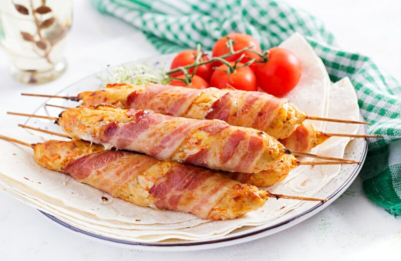 Chicken wrapped with bacon on skewers, on a white plate next to cherry tomatoes.