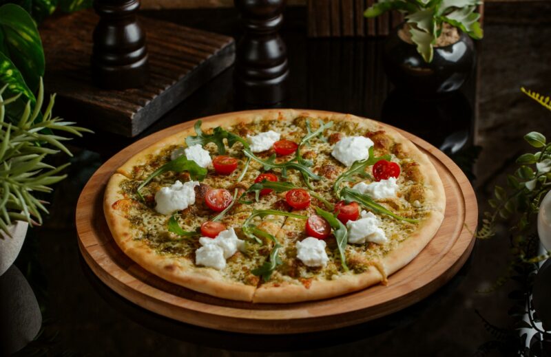 A round pizza topped with various veggies and cheese on a wooden plate in front of a dark background.