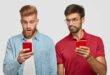 Serious beared male peeks at screen of friend`s smart phone, tries to read message, use modern technologies and high speed internet connection. Stunned red haired hipster uses cellular indoor