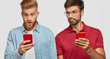 Serious beared male peeks at screen of friend`s smart phone, tries to read message, use modern technologies and high speed internet connection. Stunned red haired hipster uses cellular indoor