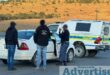 Three men appearing to be law enforcement are pictured standing next to one normal sedan and a police van at an intersection.