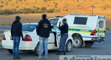 Three men appearing to be law enforcement are pictured standing next to one normal sedan and a police van at an intersection.