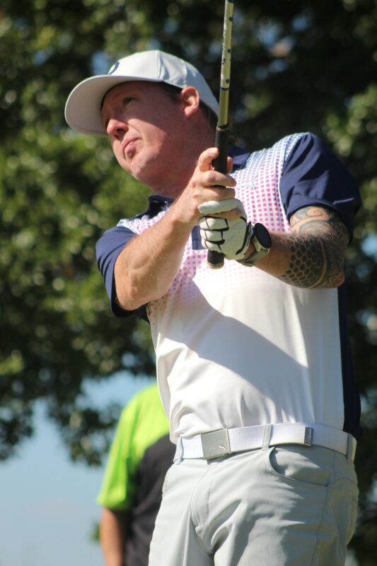 A golfer wearing a blue, purple and white T-Shirt, grey pants and a white cap is photographed teeing off on a golf coarse.