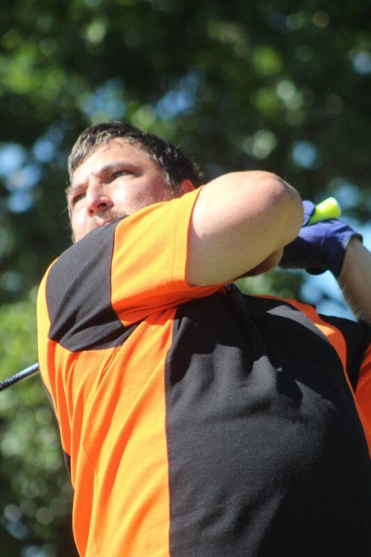 A golfer wearing orange and black attire is photographed teeing off.