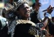 A zulu man dresses in formal Zulu cultural attire stands in front of a crowd of protestors.