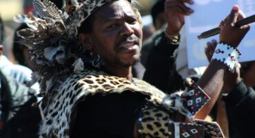 A zulu man dresses in formal Zulu cultural attire stands in front of a crowd of protestors.