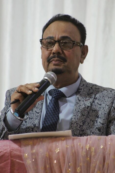 A portrait photo taken of a man in neat attire talking on a micrphone during a formal event.