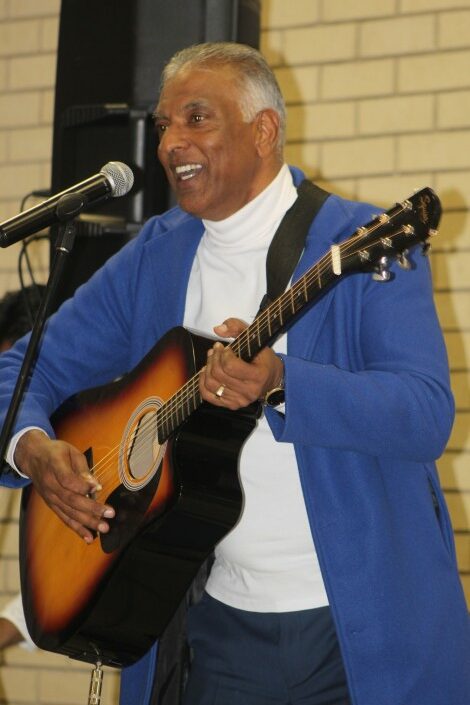 A portrait photo taken of a man wearing a white shirt and blue blazer playing the guitar.