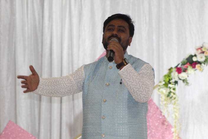 A photo of a man singing during a formal event in front of a white and pi nk backdrop.