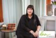 A woman dressed in black attire is sitting in her office posing for a photo. Neat and clean background setting with a chair, desk and books with a green DBM logo.