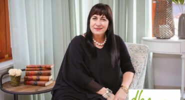 A woman dressed in black attire is sitting in her office posing for a photo. Neat and clean background setting with a chair, desk and books with a green DBM logo.