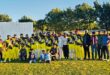 A photo of a cricket team dressed in yellow posing with supporters outside on a cricket pitch.