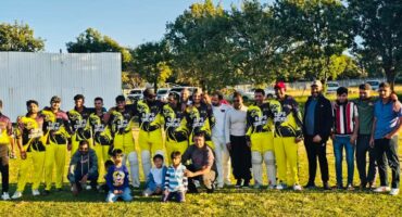 A photo of a cricket team dressed in yellow posing with supporters outside on a cricket pitch.