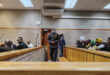 A male is seen wearing a mask entering a court room dock. Multiple people are pictured around him seated in the court room.