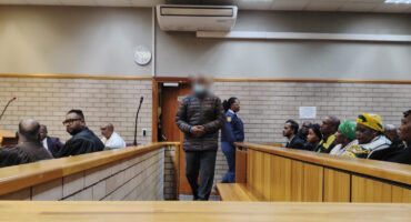 A male is seen wearing a mask entering a court room dock. Multiple people are pictured around him seated in the court room.