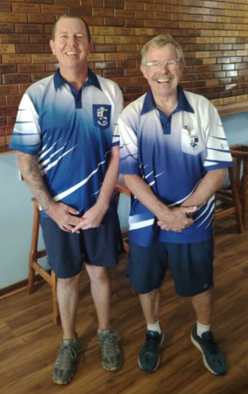 Two bowls players wearing blue and white bowls attire and posing in front of a brown, brick wall.