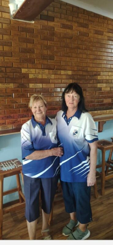 Two bowls players wearing blue and white bowls attire and posing in front of a brown, brick wall.
