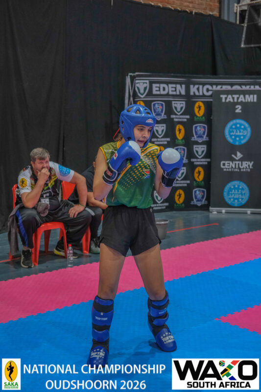 A photo capturing a young kickboxer in action while a crowd of athletes stand around supporting her on the red and blue mat.