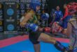 A photo capturing two young kickboxers in action while a crowd of athletes stand around supporting them on the red and blue mat.