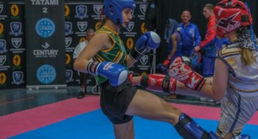 A photo capturing two young kickboxers in action while a crowd of athletes stand around supporting them on the red and blue mat.