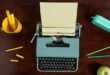A blue type writer with paper in it. A yellow cup filled with pencils and green and yellow pencils on the table.