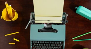 A blue type writer with paper in it. A yellow cup filled with pencils and green and yellow pencils on the table.