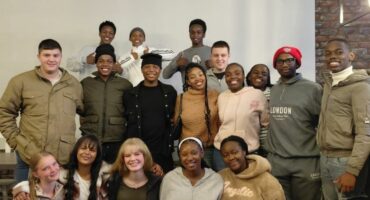 A large of young teens dressed in civvies pose for a photo in front of a white wall.