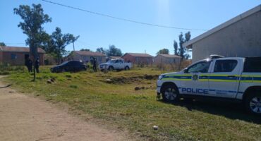 A photo taken in a rural area of a police car outside a homestead. Two other vehicles are seen in the photo with several members of the public standing outside.