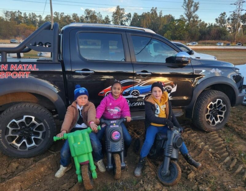 Three small children are photographed posiong on their mini plastic scooters on a racetrack.