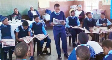 Image of learners reading from newspapers watched by educators and parents.
