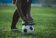 Soccer player posing with his foot on a soccer ball on a green field.