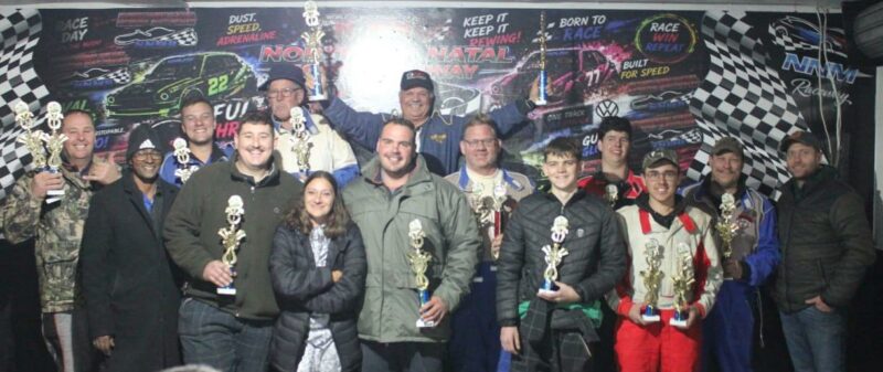 A group of Oval track winners holding their trophys pose in front of a colourful wall.