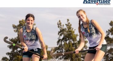 Two hockey girls are pictured holding their hockey sticks on a hockey turf behind a trophy.