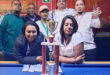 Image of group of people around a pool table with a trophy and cash prizes in the foreground.